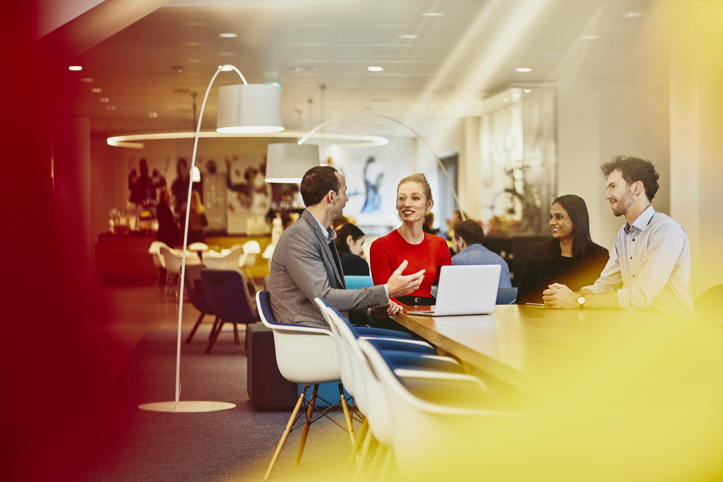 Group of colleagues sitting at a table in a meeting with a laptop, talking and smiling. Office and other people in the background. 