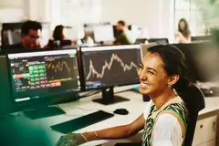 Womale smiling while sitting behind her desk, screens displaying financial information.
