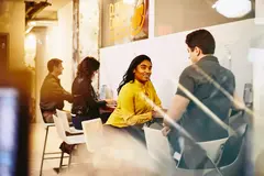 male and female having a conversation. People working on laptops in the background.