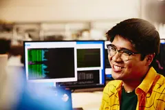 male smiling while sitting behind his desk, computer screens displaying programming code.
