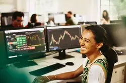 Womale smiling while sitting behind her desk, screens displaying financial information.
