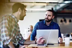 Two males talking. Both have beards. One wears glasses. One male in the background. Informal setting. Inside. Checkered shirt. Supporting color blue.
