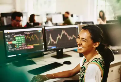 Womale smiling while sitting behind her desk, screens displaying financial information.
