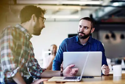 Two males talking. Both have beards. One wears glasses. One male in the background. Informal setting. Inside. Checkered shirt. Supporting color blue.