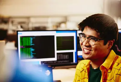 male smiling while sitting behind his desk, computer screens displaying programming code.
